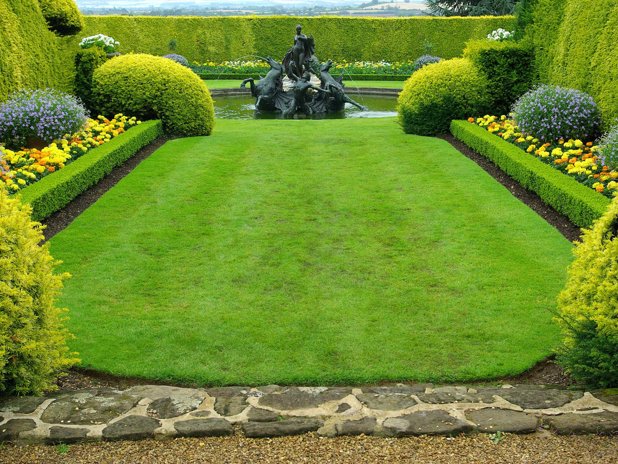 Formaler Garten mit gepflegter Rasenfläche, geraden Hecke und Brunnen mit Statue im Hintergrund
